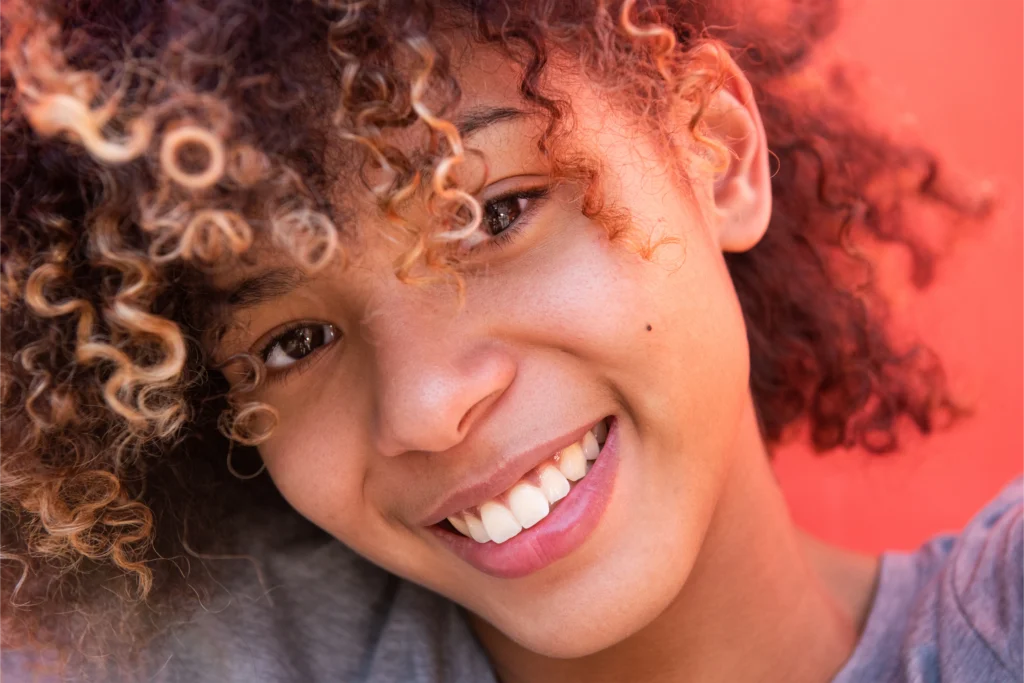 Persona joven con cabello rizado sonriendo ampliamente, mostrando dientes blancos y saludables contra un fondo de color rojo.